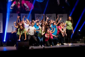 Two dozen students in colourful shirts on stage performing a musical number