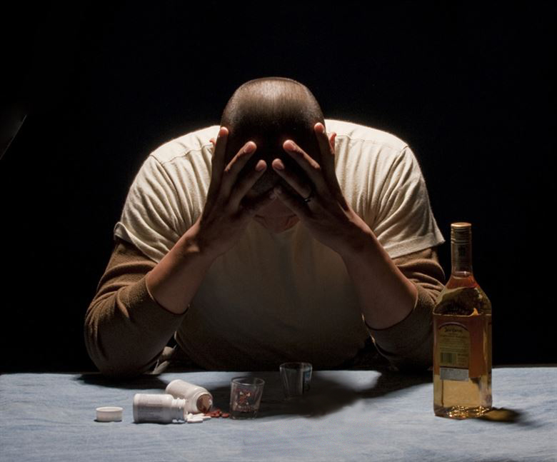 A man hangs his head in stress and holds his forehead. He is surrounded by empty glasses and pills