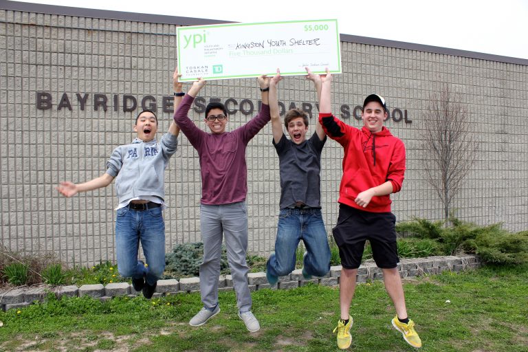 Four students jump in the air while smiling and holding a large $5000 cheque for their chosen charity.