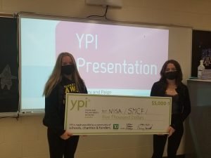 two students holding YPI giant cheque standing in front of PPT presentation